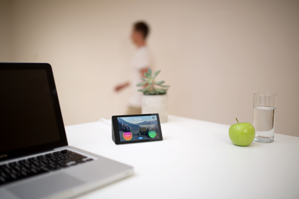 Zu sehen ist ein Schreibtisch, auf dem ein Laptop, ein Glas Wasser, ein Apfel, eine Pflanze und eine Isa steht. Die Isa zeigt den Home-Bildschirm mit dem Bewegungskreis und dem Ergonomiekreis.