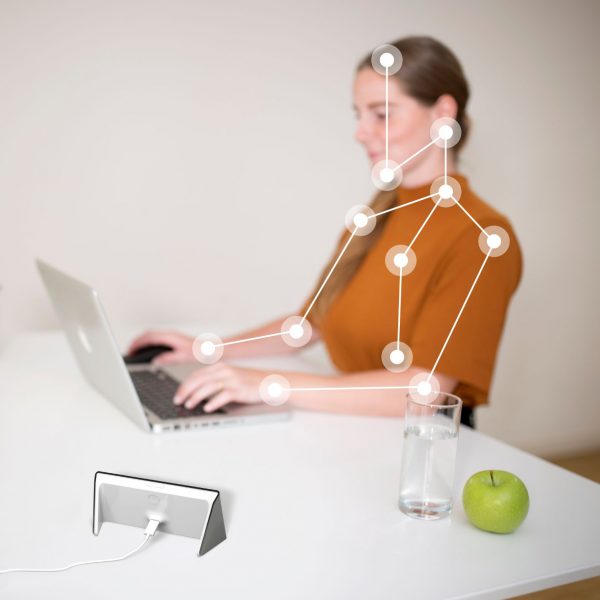 A young woman in an orange top is sitting at her desk in the office, working on her computer. An Isa stands next to her and measures the young woman's ergonomics. Dots and lines on the woman's body indicate the influence Isa has on her posture.