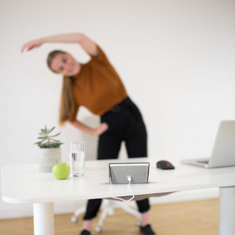 Eine Mitarbeiterin steht vor ihrem Schreibtisch, auf welchem ein Laptop, Deko, Wasser, Obst und eine Isa stehen. Auf der Isa wird ein Bewegungsübung abgespielt und die junge Frau ahmt diese Bewegungsübung nach. Sie hat einen Arm über dem Kopf und beugt sich auf eine Seite nach unten.