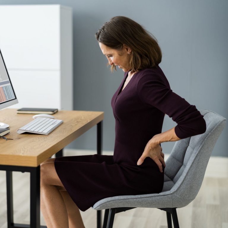 Eine Mitarbeitern sitzt am Schreibtisch in einem Businesskleid und fasst sich schmerzerfüllt an den Rücken. Sie sitzt auf einem normalen Esstischstuhl, was in der Verhältnisprävention nicht zur Ergonomie beiträgt.