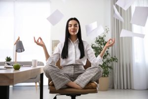Bild zeigt eine Frau, welche im Schneidersitz auf einem Bürostuhl meditiert, während um sie herum Papierblätter in der Luft schweben